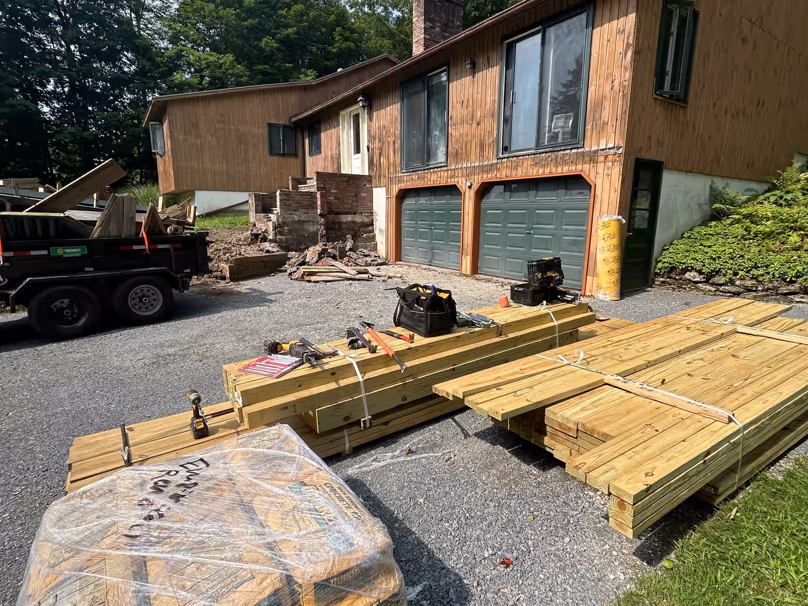 The same Vermont home after Empirical Renovations removed the deck, siding back to bare and lumber stacked in the driveway for the rebuild